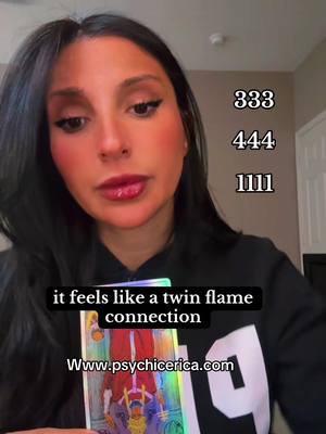 A medium shot shows a woman looking at the camera, likely recording a video. Above her head are the numbers 333, 444, and 1111 in white text. Below her, a black bar with white text reads, "it feels like a twin flame connection" and the website address, "www.psychicerica.com". The woman has dark hair, fair skin, and red lipstick. She is wearing a black hoodie, and behind her is a plain interior with a door visible in the background.