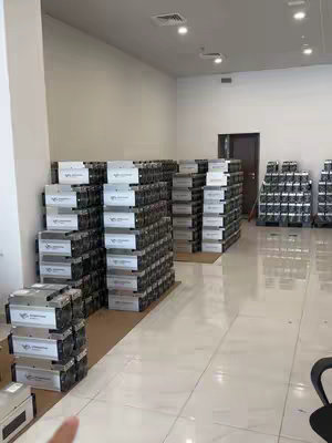 An indoor shot of a warehouse or large room filled with stacks of ASIC miners, a type of cryptocurrency mining hardware. The machines are arranged in rows, creating a sense of scale and volume. The lighting is bright and artificial, highlighting the machines and the room's polished floor. A hand is visible pointing at the miners in the foreground.