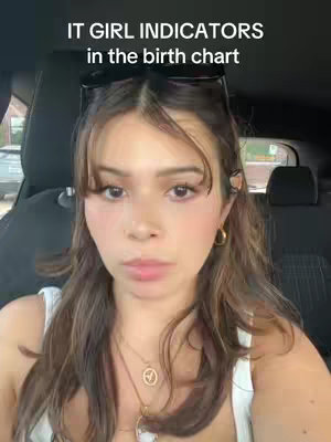 A close-up shot of a young woman with long brown hair and bangs, wearing a white top and gold jewelry. She is sitting in a car, with the car's interior visible in the background. The image has a casual, selfie-style composition.