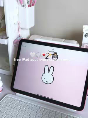 An iPad is displayed open on a table with a keyboard. On the iPad screen, a wallpaper featuring a white bunny is visible, along with the time and date in the top center. Text, 'free iPad apps every student needs,' is overlaid on the screen in white. The background includes a white shelf filled with stationary and other items, as well as a portion of the desk.