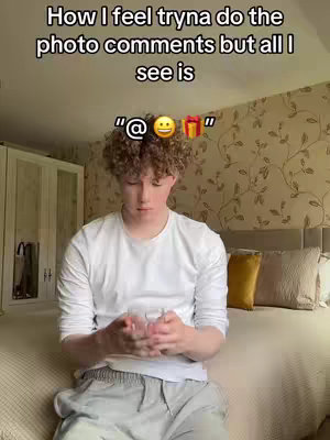 A young man is the primary focus, positioned centrally in the frame. He's wearing a white long-sleeved shirt and light-colored sweatpants, suggesting a casual indoor setting. His curly hair is neatly styled. He is looking down at something he holds in his hands while sitting on the edge of a bed. The background includes a bed with neutral-colored pillows and a floral patterned wall. The overall ambiance leans towards a casual, personal setting.