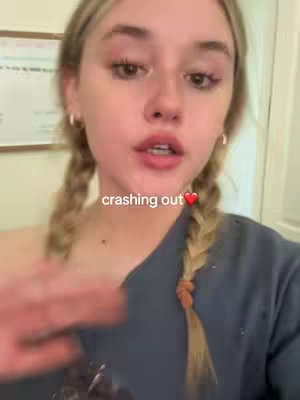 A young woman with blonde pigtails is centered in a selfie-style video. The background is slightly blurred, showing what appears to be a room with a framed item on the wall. The woman is looking towards the camera with a slightly open mouth, appearing to be speaking. The image is well-lit with natural light.