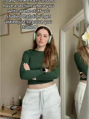 A young woman with reddish-brown hair, wearing a green long-sleeved shirt and white sweatpants, stands with her arms crossed. She's wearing headphones and a smartwatch, and the background is a simple room with a mirror and a framed picture.