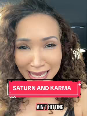A selfie-style image of a woman with dark curly hair and makeup, smiling directly at the camera. A rectangular red and black text overlay saying "Saturn and Karma" is across the midsection of the image. Below it, in smaller text, is the phrase "Ain’t Hitting". The background is blurred, suggesting an indoor setting. The overall composition is a close-up shot emphasizing the woman's expression.
