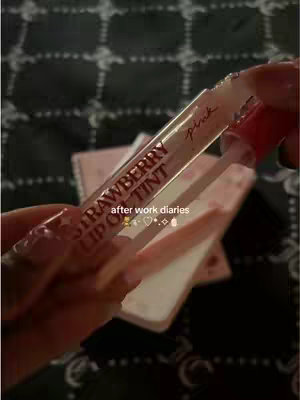 A close-up shot captures a hand holding a tube of pink strawberry lip gloss. The tube is mostly transparent, with the product visible inside and the word “pink” written across the top. The background is blurred, but suggests a dark surface. Added text says "after work diaries".