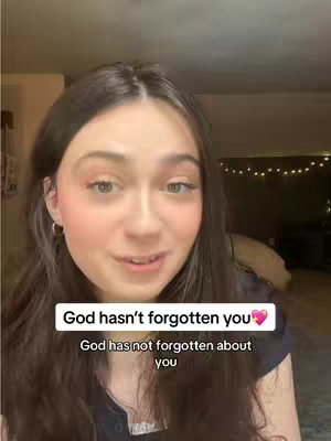 A young woman with long brown hair, wearing a dark top, is the central figure. She has fair skin, rosy cheeks, and is looking at the camera with a slight, gentle smile. The background is softly lit, with hints of a room, showcasing a blurred, warm palette. Above her head, a white rectangle contains the words "God hasn't forgotten you💖" and a slightly smaller white rectangle directly beneath it contains "God has not forgotten about you".