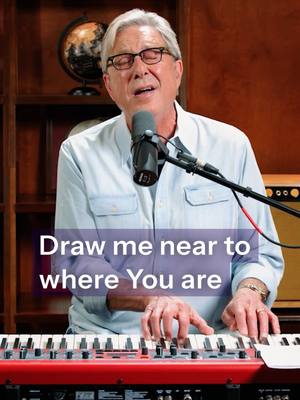 A man with gray hair and glasses is seated at a keyboard, with a microphone in front of him. Text in a purple box reads, "Draw me near to where You are". The background includes a shelf with a globe and some wooden elements.