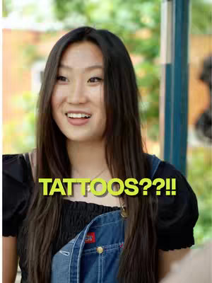 A young asian woman smiles broadly, looking directly at the camera. She has long, dark hair and wears a black top under blue denim overalls. The words 'TATTOOS??!!' are written in large, yellow, bold text across the image.