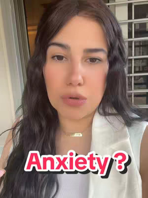 A woman with dark wavy hair looks directly at the camera, with a soft, natural glow. She wears a white blazer over a white top and a small gold necklace. The background is slightly blurred, suggesting an indoor setting with a light-colored wall and a glimpse of an outdoor area with a metal railing. The text overlay 'Anxiety?' appears in a bold, red-outlined font, positioned over the lower part of the image..