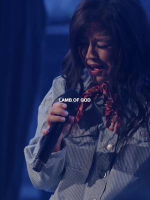 The cover image features a woman with dark hair, illuminated from the top, singing into a microphone. She wears a striped button-down shirt, and a printed scarf around her neck. The text "LAMB OF GOD" is overlaid.