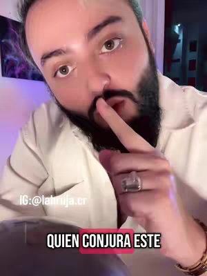 A medium shot features a man with a dark beard and short dark hair. He is wearing a white shirt and has a large silver ring on his right hand. He is bringing his index finger to his lips in a shushing gesture. Overlay text in a black box with a pink outline reads "QUIEN CONJURA ESTE". The background is slightly blurred, showing a framed artwork of a galaxy.
