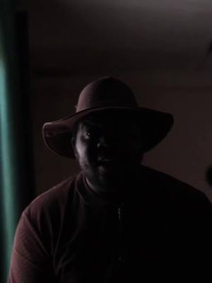 The image is a low-key portrait featuring a person wearing a hat, with the face mostly obscured by shadow. The lighting is dim, creating a strong contrast that emphasizes the silhouette of the person and the hat. The person's expression and other details are concealed due to the reduced lighting.