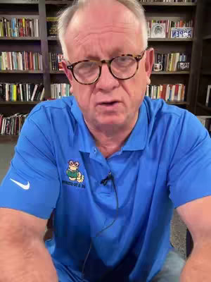 A medium shot of a man with glasses and a blue shirt, speaking into the camera. A bookshelf fills the background. The man has fair skin and short hair.