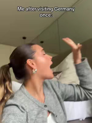 A woman with her hair in a ponytail is shown from the side, yelling with a surprised expression. The background is blurry, with a glimpse of a bed and a ceiling. Text overlay reads "Me after visiting Germany once:"