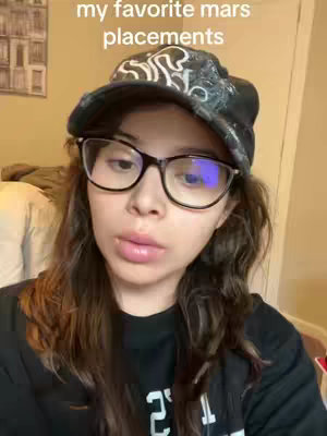 A woman with long brown hair wearing glasses and a black cap smiles slightly at the camera indoors. Text overlayed on the top of the screen reads 'my favorite mars placements' in white font.