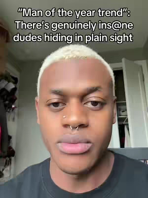 A selfie-style shot of a young Black man, with a close-up focus on his face. He has short, light-colored hair with a styled fringe. He wears a nose ring and a black shirt. The background is slightly blurred, showing a glimpse of a light-colored wall and a door.