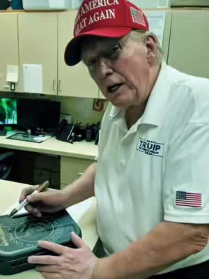 Donald Trump is shown in a room, wearing a red 'Make America Great Again' cap and a white polo shirt with the 'TRUIP' logo. He's holding a pen and a dark object and appears to be signing it. The background includes cabinets and a computer.