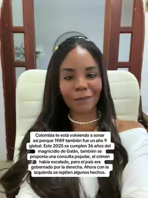 A woman with long dark hair poses for a selfie, seated in a well-lit room.  She wears a white top, and a string of pearls.  Text overlays her lower half.