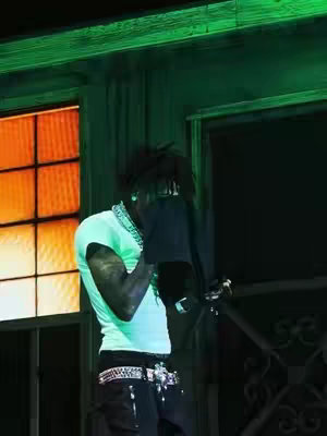 The cover image shows a person, presumably the content creator, partially obscured by a black object, possibly a piece of clothing, in what appears to be an outdoor setting. The person is dressed in a light green t-shirt, adorned with jewelry around their neck, and black pants with a bedazzled belt. The background features a window setting with an orange glow on one side and a dark area on the other, both with green overtones, suggesting a specific lighting or time of day.
