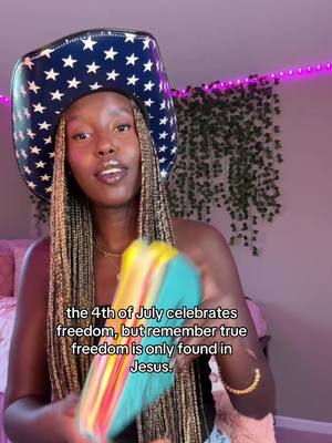 A young Black woman wearing a cowboy hat with stars poses for a selfie, holding a deck of colorful cards in her hands. Behind her, a wall features hanging vines and glowing pink lights, suggesting an indoor setting. Text overlayed at the bottom of the image reads: "the 4th of July celebrates freedom, but remember true freedom is only found in Jesus."