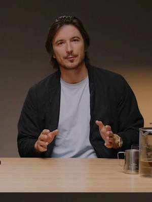 A man with brown hair and a black jacket is sitting at a table. He is speaking with his hands gesturing upward, and there is a cup on the table behind him.