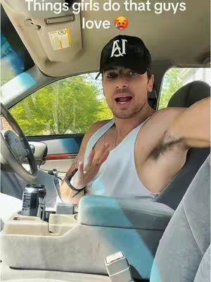 A man is sitting in the driver's seat of a car and talking. The background shows trees and greenery outside the car window. The man is wearing a baseball cap and a tank top. Both of his arms are in the image, while he touches his face with his right hand and gestures with his left hand.