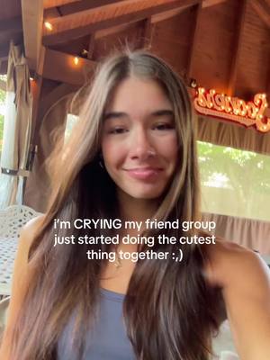 A young woman with long brown hair is facing the camera, smiling slightly. She's wearing a blue top and a small necklace. The background shows a wooden structure and some foliage, suggesting an outdoor setting. Text overlays the image.