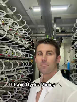 A man stands in front of a complex setup of white tubes and machinery, likely part of hydro mining operations. The background industrial setting contrasts with the man's approachable attire.
