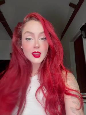 A woman with long, vibrant red hair is the central focus. Her face is framed by the flowing hair, and she is looking directly at the camera with open lips and visible red lipstick. She is wearing a white top, and behind her, the background includes dark wooden beams and the top of a door frame.