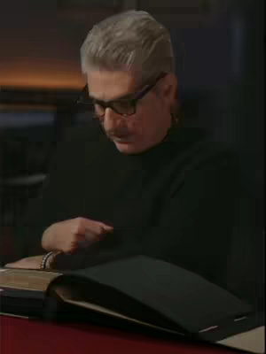 A medium shot shows Michael Imperioli looking down with his hands clasped in front of him on a book laid open on a table. He has short gray hair, glasses, and a mustache and is wearing black clothing. The lighting is dim, and the background is blurred.