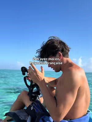 A shirtless man sits on a boat or watercraft, holding a diving mask. The background shows clear blue water under a bright sky.
