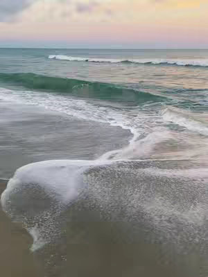 The image shows a serene ocean scene at dawn or dusk, with a predominantly calm ocean transitioning to a sandy beach in the foreground. The sky transitions from a soft pink and yellow at the horizon to a muted blue higher up. Gentle waves are breaking on the shore, and the water appears clear and calm. The overall composition evokes a sense of peace and tranquility.