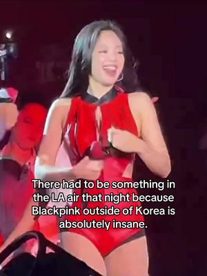 A woman with long black hair, wearing a red outfit, is on a stage. She is smiling broadly and appears to be actively engaging. The background is dark with stage lighting illuminating her.
