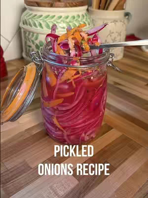 A jar of pickled onions takes center stage, resting on a wooden surface. The onions, bright pink, are tightly packed within the clear glass jar, and topped with chopped vegetables. A spoon rests at the rim of the jar. Additional background elements include other jars.