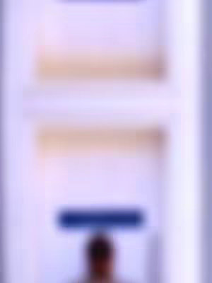 The image is a blurred, abstract composition primarily in shades of light purple and beige. The top part of the image appears to be a series of horizontal, slightly blurred lines suggestive of architectural elements or shelves, with light beige tones. In the center, there's a darker, rectangular shape, possibly a sign or a structural detail, in a muted blue color. Below this, there is the blurry outline of a person, indicating that it is a photo of a person in the background. The overall composition gives a sense of depth and a soft, unfocused aesthetic.