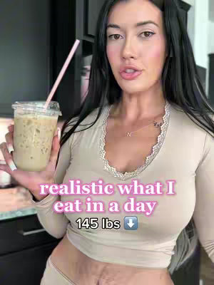 A woman with long dark hair is holding a coffee drink, looking towards and speaking to the camera. She is wearing a light-colored top with lace detail, and the words 'realistic what I eat in a day' and '145 lbs' are superimposed in a pink stylized font. The background seems to be a kitchen setting with stainless steel appliances.