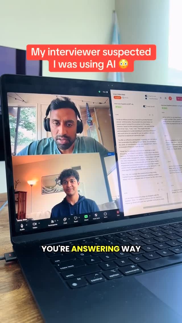 A laptop screen displaying two video call windows. The top window features a man with dark hair wearing headphones. The lower window displays the same man earlier in the video call with curly hair. A colored bar above the laptop screen reads 'My interviewer suspected, I was using AI 🤔'. At the bottom a text overlay reads 'YOU'RE ANSWERING WAY'.
