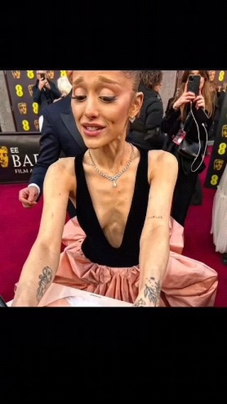 The image shows Ariana Grande in a candid moment, likely at an event. She is positioned from a slightly low angle, with a focus on her upper body and face. She's wearing a black top with a plunging neckline and a light pink, voluminous skirt. A diamond necklace is visible. Her expression appears to be one of slight surprise or concern., with visible tattoos on her arms. The background is blurred. The overall composition suggests a close-up, capturing a personal moment in a public setting.