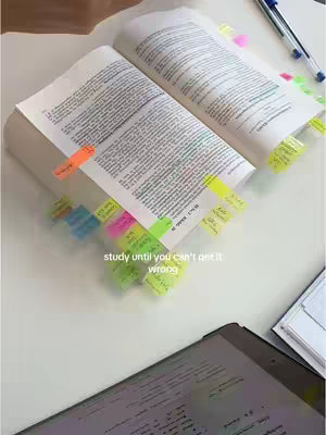 An overhead shot of an open book with numerous sticky notes marking pages. A tablet lies in the foreground, and a pen is casually placed on the surface. The colors are muted, with the white surface contrasting the book's pages and the colorful sticky notes.