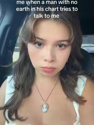A close-up shot features a young woman with brown hair and light skin, wearing a white top and a cameo necklace. She has a slightly displeased expression. The background is blurred, suggesting she is in a car.