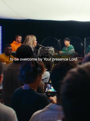 The cover image features a live performance in a venue, likely a church or concert hall. In the foreground, a crowd is partially visible, with several people holding phones. On the stage, a person with blonde hair is singing into a microphone. Behind her, other musicians and audio equipment are present, and the background seems to be a dark stage setting. The text overlay reads: "to be overcome by Your presence Lord". The lighting is bright, focused on the performers, and a soft white ceiling is visible above.