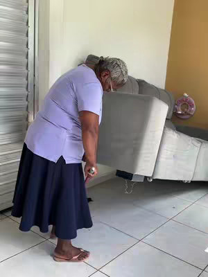 An elderly woman is seen indoors, bending over to seemingly lift or adjust a large, gray couch cushion. She is wearing a light purple short-sleeved top and a long, dark blue skirt, paired with pink flip-flops. She is facing downward, with her back to the camera, her gray hair pulled into a bun. The background features a white wall, a glimpse of a door and some decorative elements. The composition focuses on her activity, capturing a moment of everyday life.
