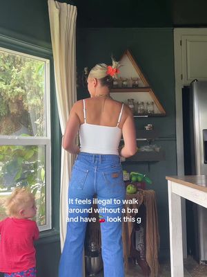 A woman stands with her back to the camera, wearing a white tank top and high-waisted jeans. She has blonde hair with a flower accessory. A young child is visible in the foreground, looking at the woman. The background includes a green-painted wall, a window, and kitchen elements.