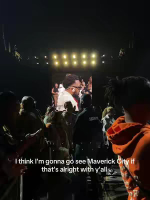 The cover image captures a concert scene. The foreground is filled with the backs of numerous people, likely audience members, facing the stage. In the middle ground, a large screen displays a person on stage. The background is dark, and the lighting suggests a concert setting.