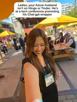 A selfie-style shot with a woman smiling and extending her arm towards the camera, taken at an outdoor event. She's wearing a black top, a badge, and a lanyard. The background includes yellow umbrellas, tables, and people, suggesting a tech conference or similar event.