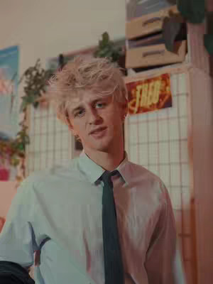 A young, fair-skinned man in a light blue button-down shirt and a dark tie stands in front of a cluttered background. His light blonde hair is styled upwards, and he has a slight smile. Behind him, a grid-like structure with various items and a sign that reads 'FRED' hints toward a room's interior.