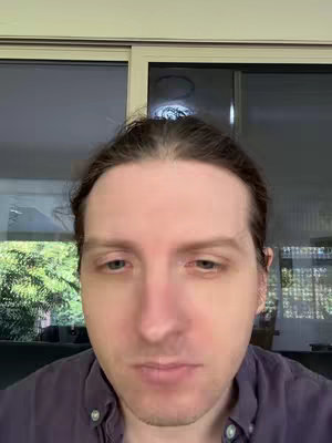 A close-up shot of a man's face, centered in the frame. He has light skin, brown hair with a receding hairline, and light eyes. He wears a dark purple button-down shirt. The background shows a blurred window and an interior setting. The overall composition has a natural lighting.