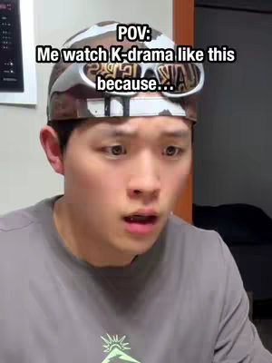 A selfie-style shot featuring a young man with dark hair and fair skin. He wears a camouflage hat with sunglasses, and a neutral-colored shirt. The background is plain. He has a surprised expression with his mouth slightly open as if in astonishment. Text overlay on the image reads, "POV: Me watch K-drama like this because..."