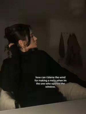 A dimly lit image features a woman in profile, leaning against what appears to be a headboard or wall. She wears a black sweater, with dark hair pulled back, and delicate earrings. Text overlayed reads "how can i blame the wind for making a mess when im the one who opened the window."
