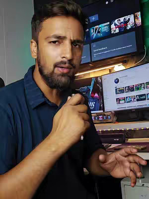 A man in a navy blue collared shirt is holding a microphone and looking towards the viewer with a slightly concerned expression. Behind him, a television displays the interface of a streaming service, and to the right, a computer monitor is also visible. The background includes a desk and other electronics.